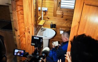 Un grupo de personas observa atentamente la instalación de un sistema Smart Water en un ambiente interior de madera, capturando el momento en que la tecnología de conservación del agua se integra en la vida cotidiana