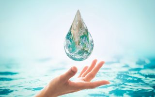 Una mano sostiene cuidadosamente una gota de agua gigante transparente, con el planeta Tierra visible en su interior, sobre un fondo azul claro que simboliza la importancia de la conservación del agua para nuestro mundo.