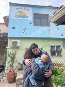 Propietario de la Casa Rural Lago de Anna abrazando a una familia frente a la fachada decorada con murales y un cartel identificativo.