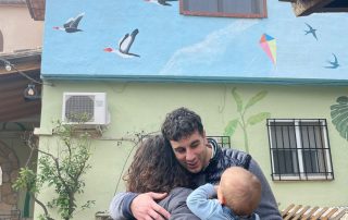 Propietario de la Casa Rural Lago de Anna abrazando a una familia frente a la fachada decorada con murales y un cartel identificativo.